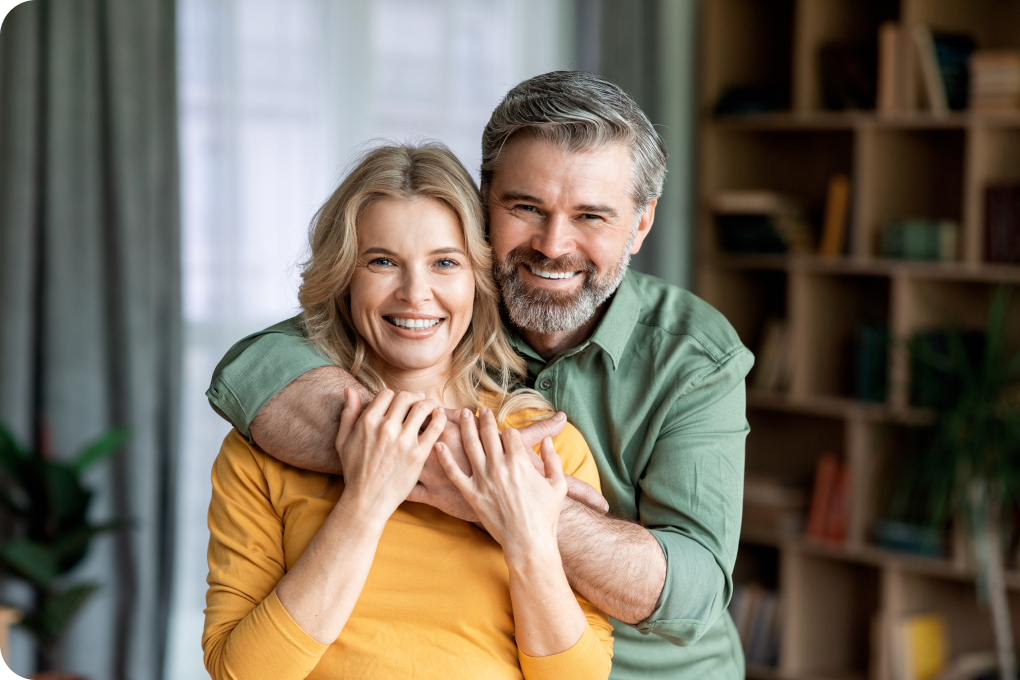 Homem e mulher sorridentes abraçados olhando para frente. Eles vestem camisas de mangas compridas. O ambiente é uma sala com uma prateleira de livros no fundo. 